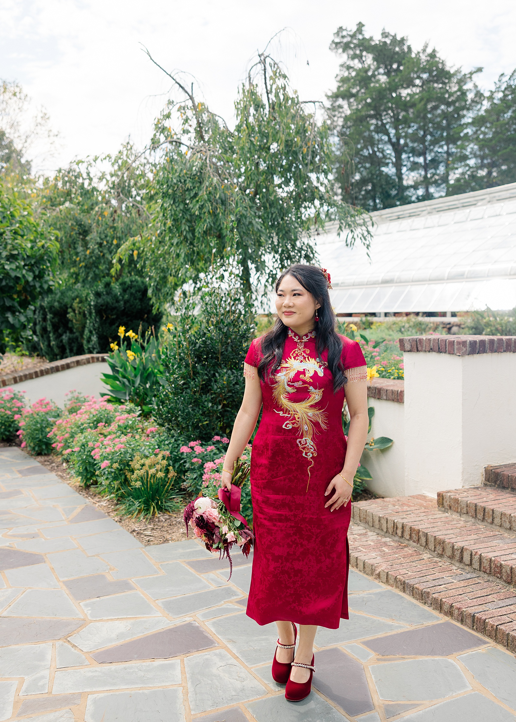 A giggling bride in a red dress holds her bouquet while walking by a greenhouse in a garden