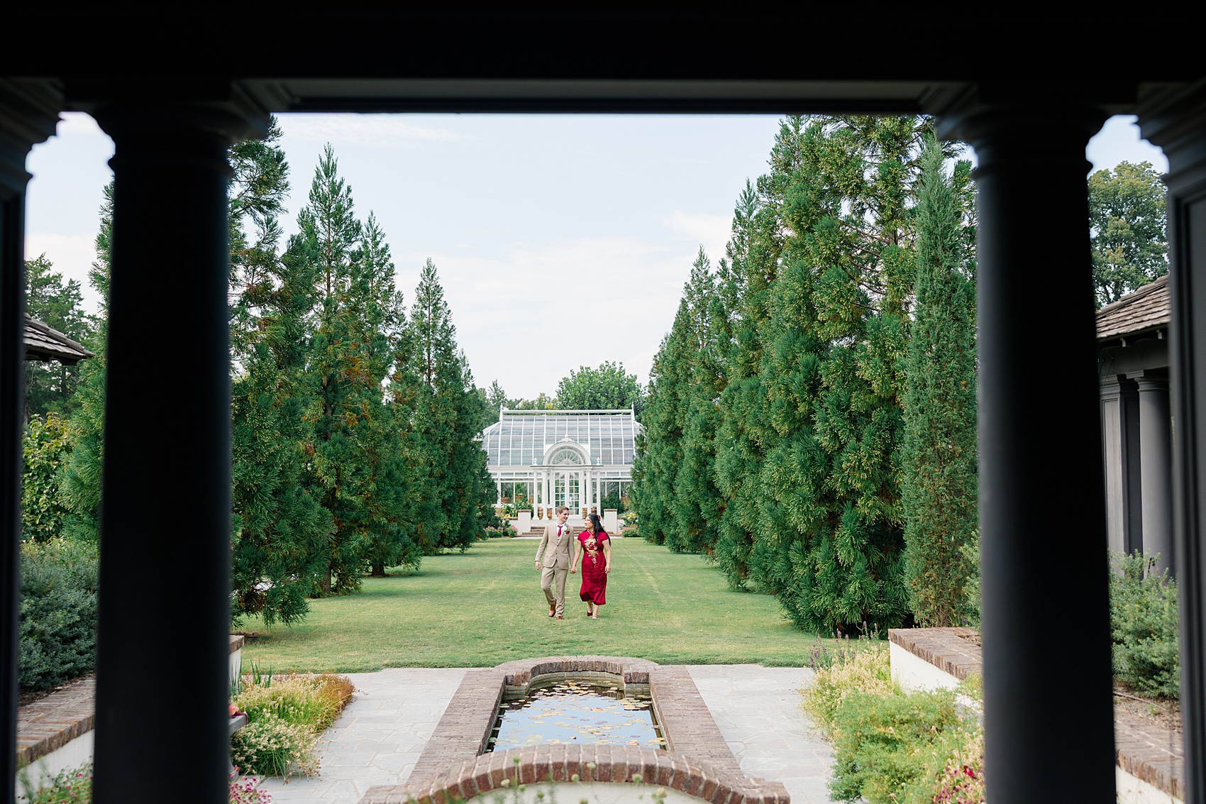 A bride in red walks holding hands with her groom in tan through the gardens before the barn at reynolda village wedding reception